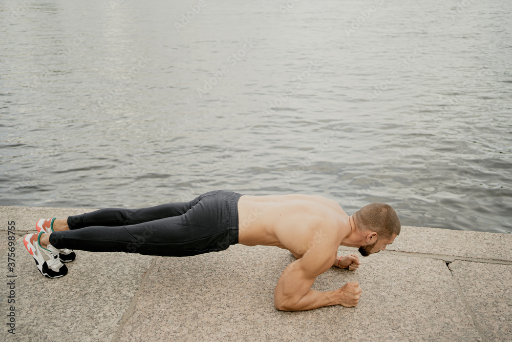 athletic dark-haired man doing a plank exercise on the street near the water, tense looking ahead. short haircut brunette barbershop. Caucasian appearance athletic dark-haired man doing a plank exercise on the street near the water, tense looking ahead. short haircut brunette barbershop. Caucasian appearance