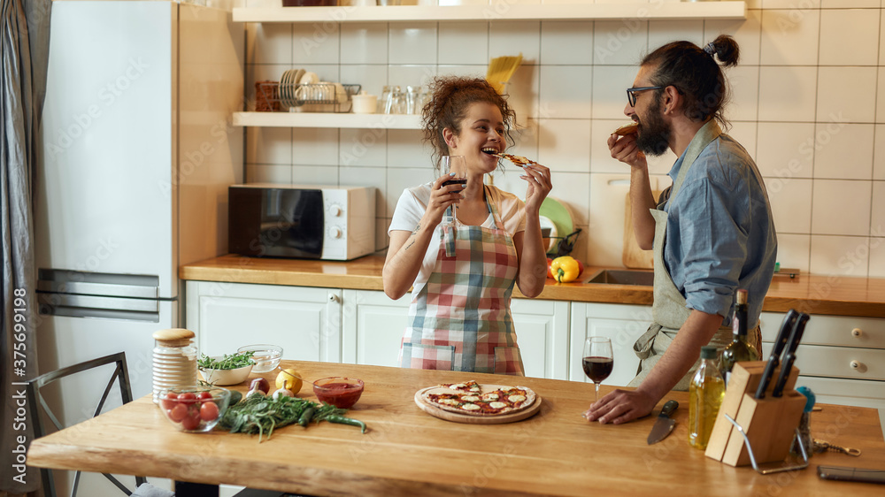 Enjoy it. Young man and woman in apron eating freshly baked pizza and drinking wine while standing in the kitchen. Love, relationships concept