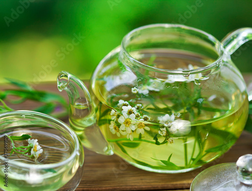 Herbal tea with achillea salicifolia flowers in a glass teapot and glass thermo cup on a green bokeh background.