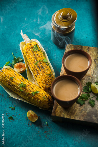 Top view of brewed hot milk tea with roasted grilled corn on blue background. Delicious Grilled Corncobs and Two Cups of Chai, an ideal Indian monsoon brunch good for boosting immunity and health.