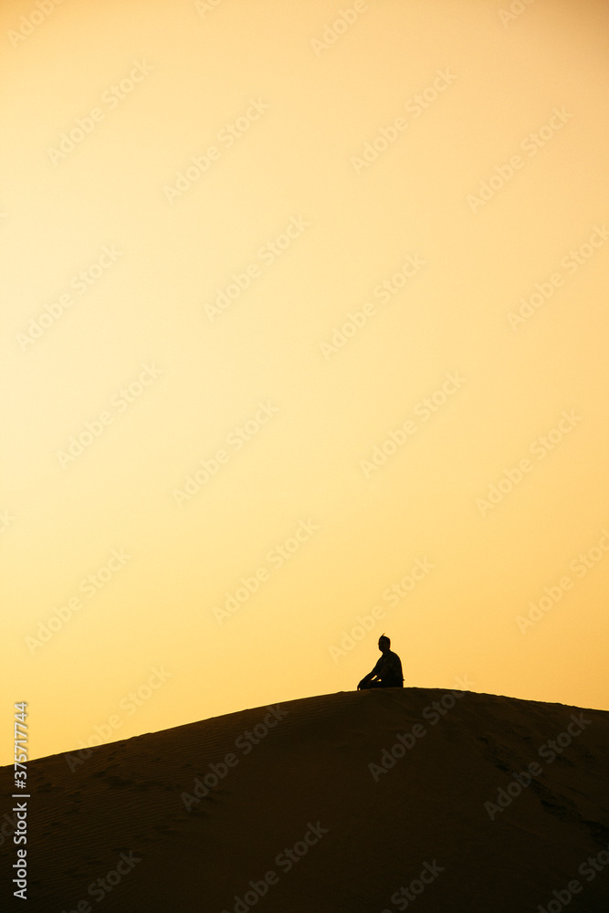 A silhouetted man sitting on top of a sand dune, contemplating. A silhouetted man sitting on top of a sand dune, contemplating.