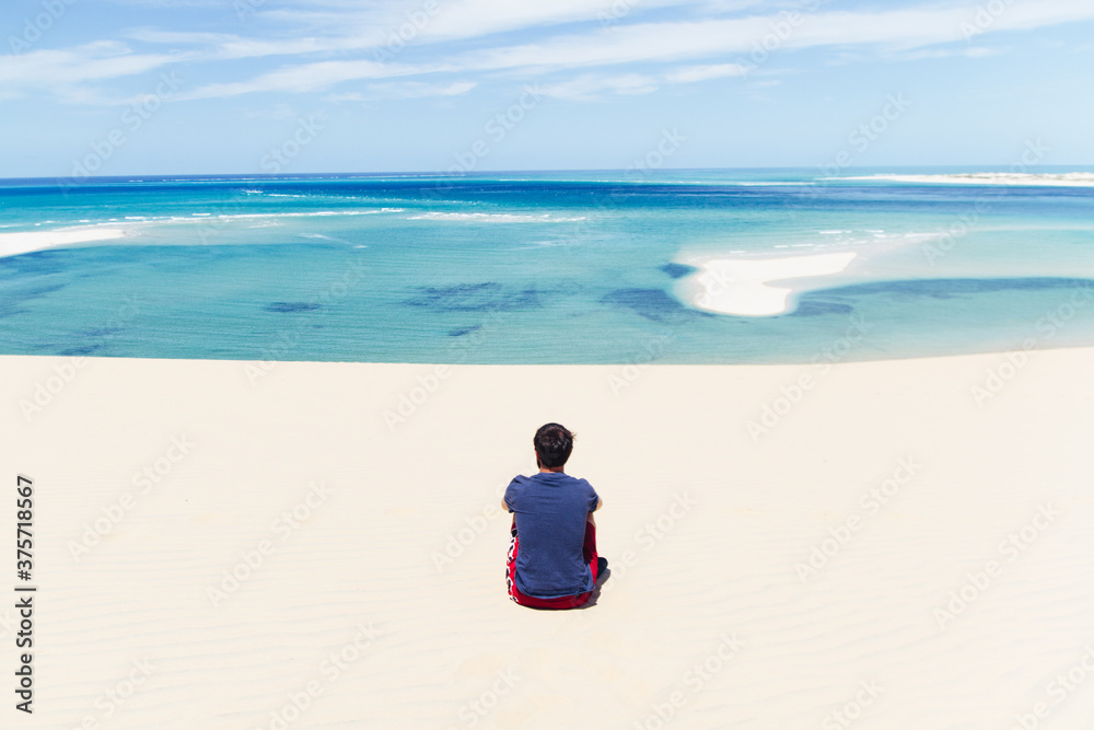 Young man sitting down on the sand looking at the ocean on an exotic tropical beach with turquoise water Young man sitting down on the sand looking at the ocean on an exotic tropical beach with turquoise water