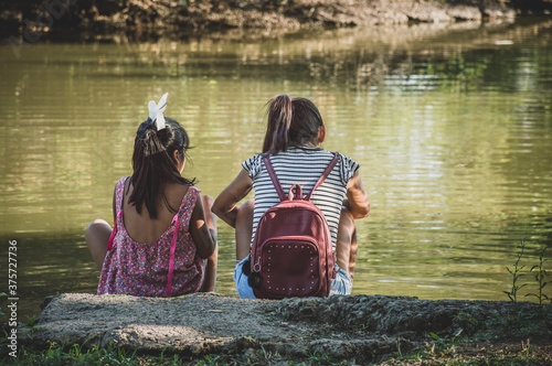 Obraz na płótnie Unrecognizable two little girls sitting by a lake. Concept of childhood. w salonie