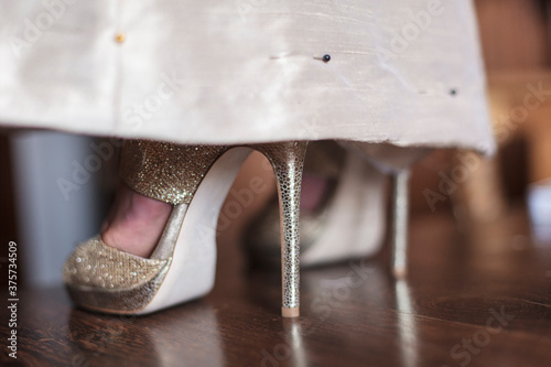 A women's feet in sexy, gold shoes peak out from the hemline of a gold dress that is pinned up. photo