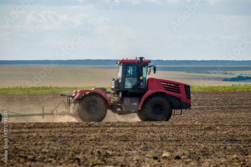 A red farm tractor in a cloud of dust cultivates the soil in the field with a cultivator after harvest. Summer sunny day. Fertile land. Modern agricultural machinery. Copy space. High quality photo