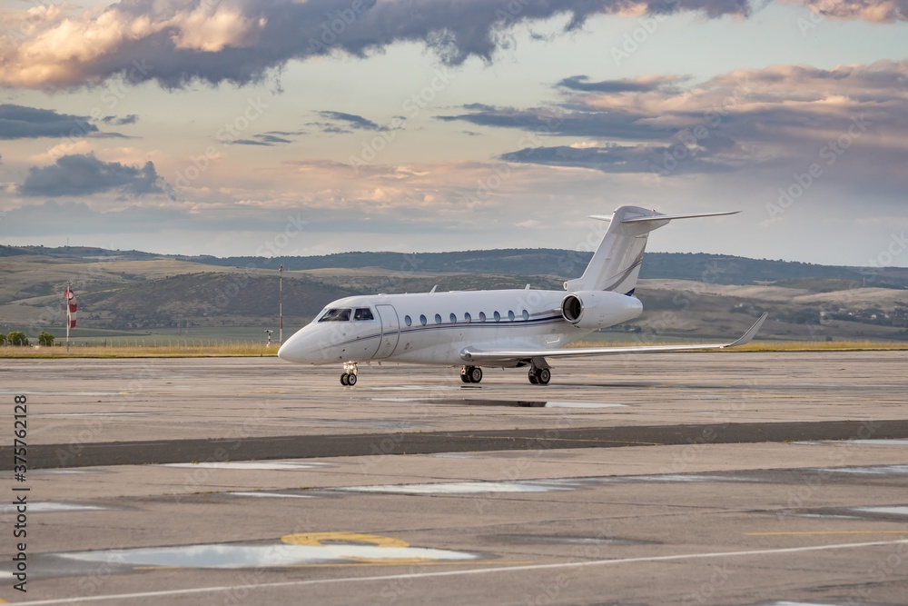 White small private twin-engine business jet on airport apron. Attractive landscape dramatic grey orange clouds, puddles after rain weather. Modern technology in fast transportation, business travel.