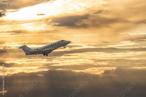 White small private turbofan-powered twin-engine business jet airplane flying in air after takeoff from airport. Dramatic orange grey cloudy sky at sunset after rain storm, dynamic weather conditions.