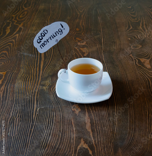 A white cup with green tea stands on the table surface. There is a tablet with an inscription in ink to the pen. Good morning mood. Background with wood texture. Hot fresh drink.