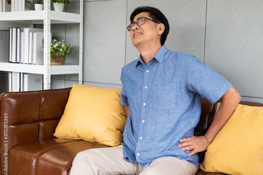 Indoor shot of senior Asian men sit on the sofa couch and touch his back and suffering from back pain, aching muscles, bones or lumbago. Elderly health problem concept.