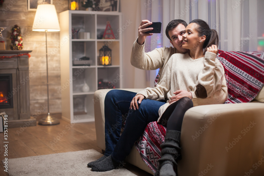 Obraz premium Smiling couple taking a selfie celebrating christmas day sitting on couch.