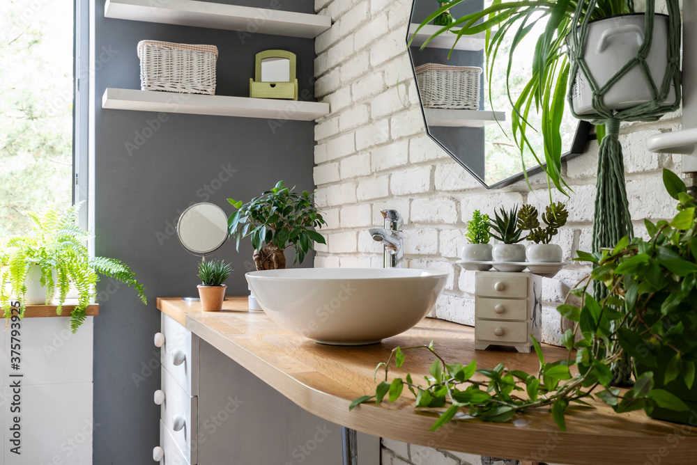 Stylish bathroom with white brick wall, mirror, green plants, washbasin and wooden furniture. Bright interior of spa in houseplant. 