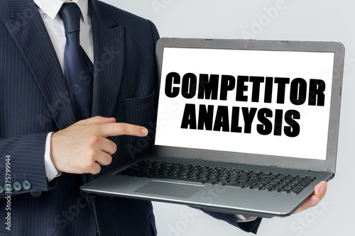 A businessman holds a laptop in his hands and shows on the screen with the text - COMPETITOR ANALYSIS