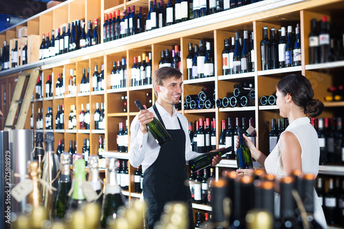 Seller wearing apron helping to buy bottle of wine to woman customer in wine store