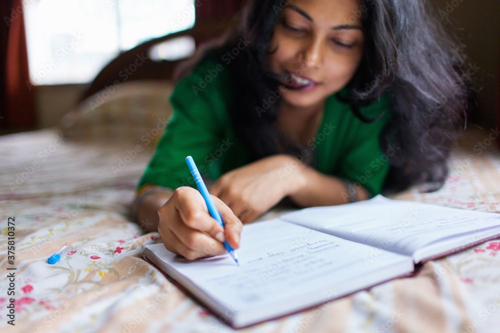 Young woman writing on her diary while lying on bed Young woman writing on her diary while lying on bed