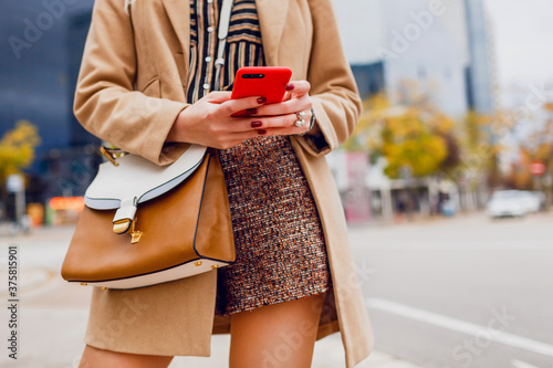 Woman hands with mobile phone. Stylish girl in beige coat chatting. Modern city background.