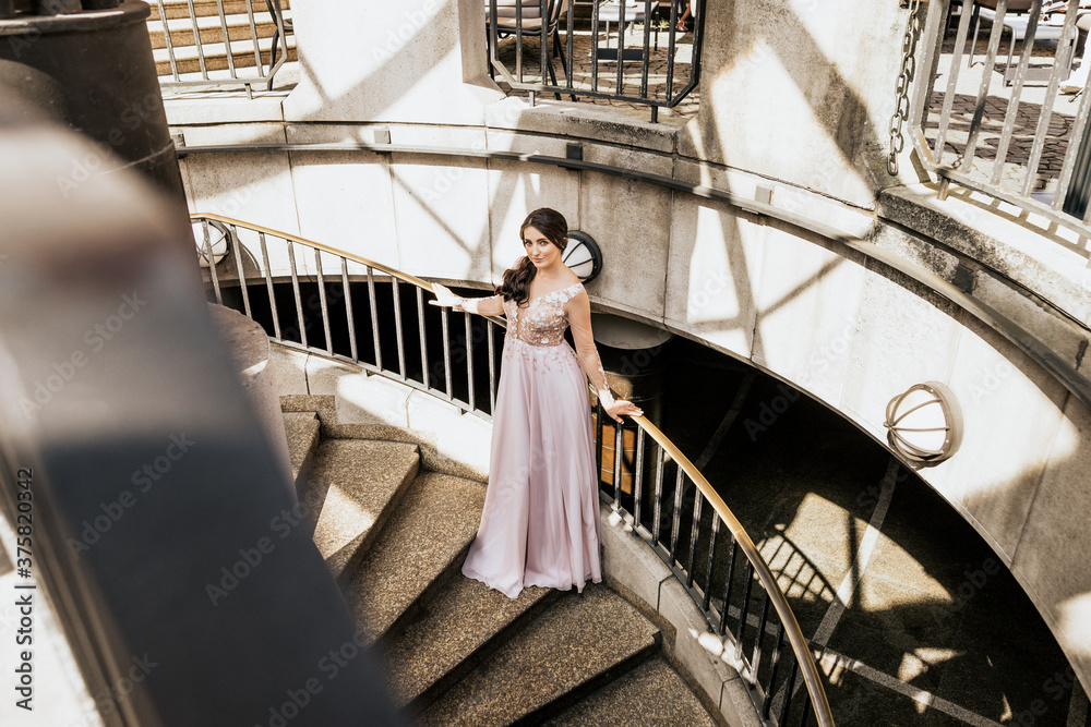 Beautiful attractive woman stand on stairs in pink dress and look in camera. Young woman inside building