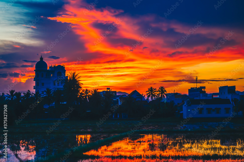 Fiery dark orange red yellow purple colorful natural bright dramatic sunset sky. Evening cloudscape reflected in water, background building in shadow, ducks floating in rice field. Design wallpaper