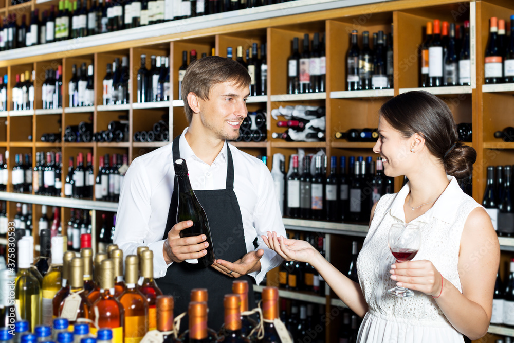 Young man seller wearing uniform giving to woman customer try glass of wine in wine house