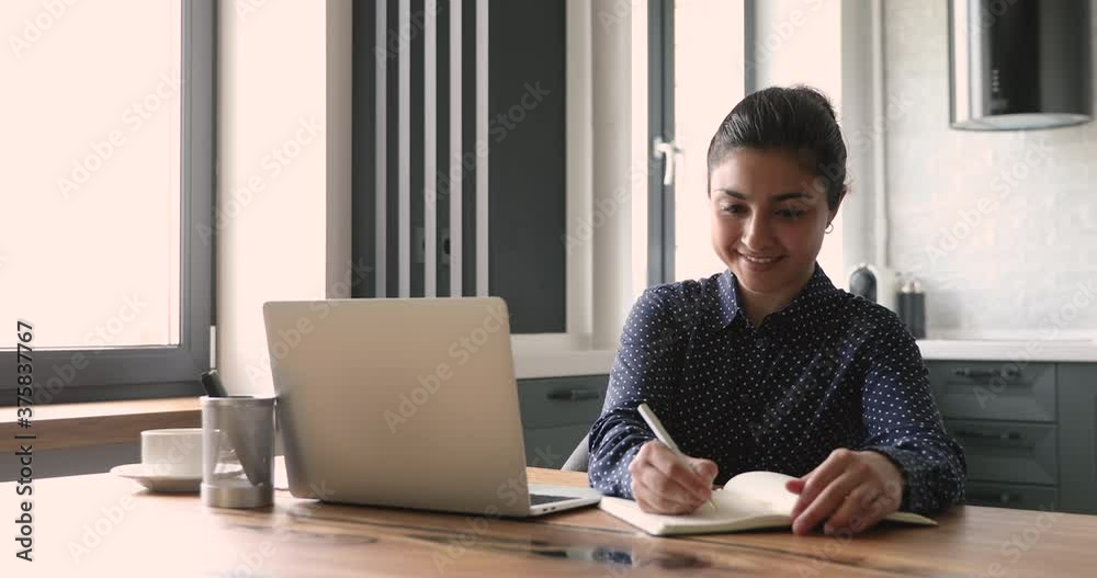 Smiling indian business lady working from home office sit at desk use laptop search information on internet writing down it in organizer agenda. Self-education, notebook personal planner usage concept Smiling indian business lady working from home office sit at desk use laptop search information on internet writing down it in organizer agenda. Self-education, notebook personal planner usage concept