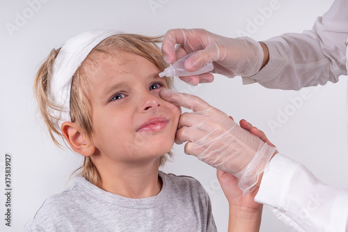 Female hands optometrist wearing transparent medical gloves putting eye drop patient little blonde boy eyes in ophthalmology clinic. Sick look. White background.