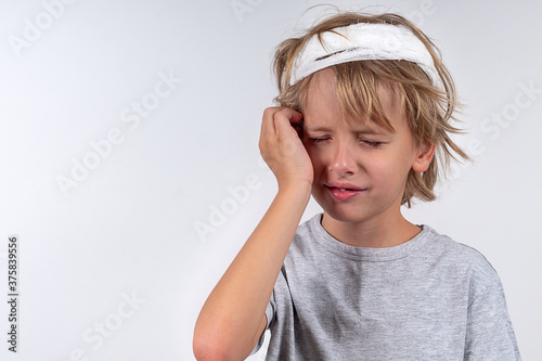 Portrait young caucasian cute boy blond hair with trauma injury and bandage head. Isolated on white background. Boy holds his head with his hand.