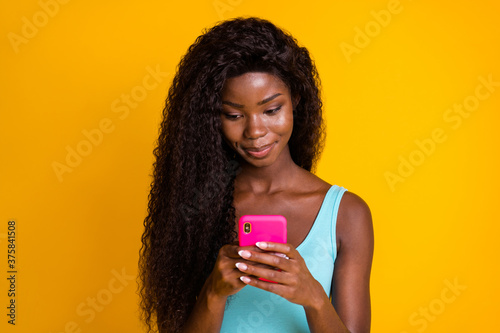 Photo portrait of young african american girl holding pink phone in both hands wearing blue tank-top isolated on bright yellow colored background