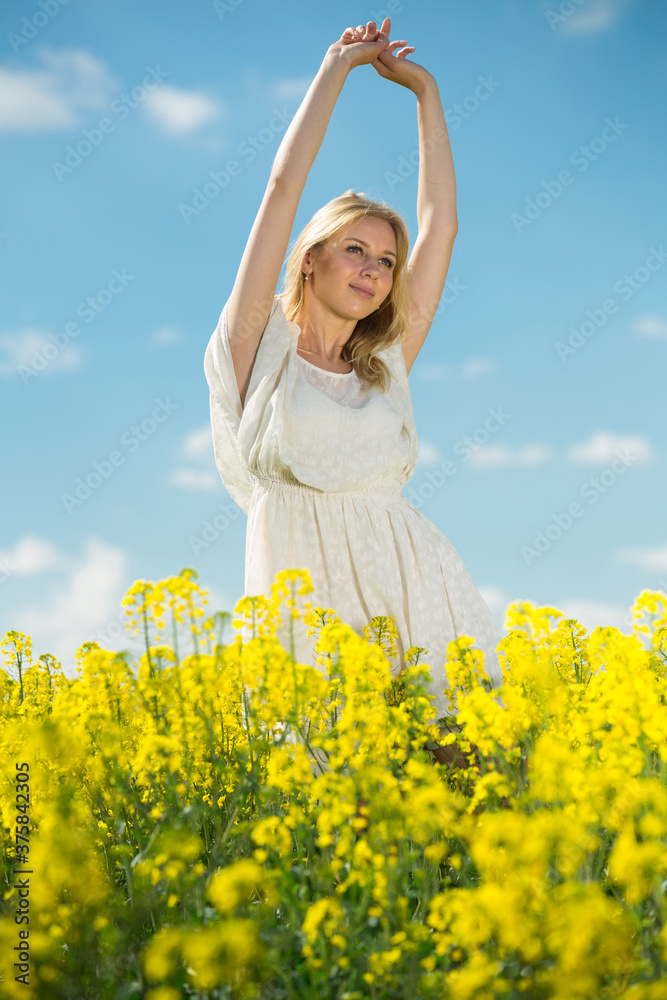 Fototapeta premium Happy girl stretch oneself in rape seed flowers field posing in white dress at sunny day outdoor