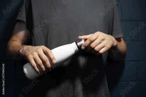 Close-up of male hands, openes steel thermo water bottle of white color, on the background of black brick wall. photo