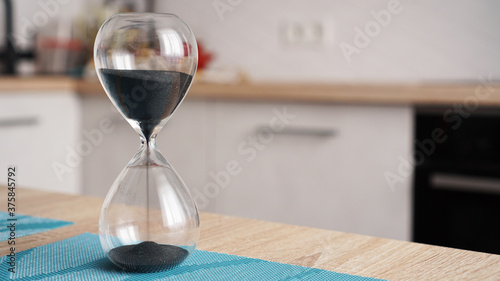 Closeup of hourglass on a white wooden table with defocus kitchen on background. Concept of time to cook.