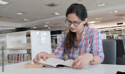 Obraz na plátně Photo of beautiful and intelligent young girl with eyeglasses studying hard in public library for upcoming exams