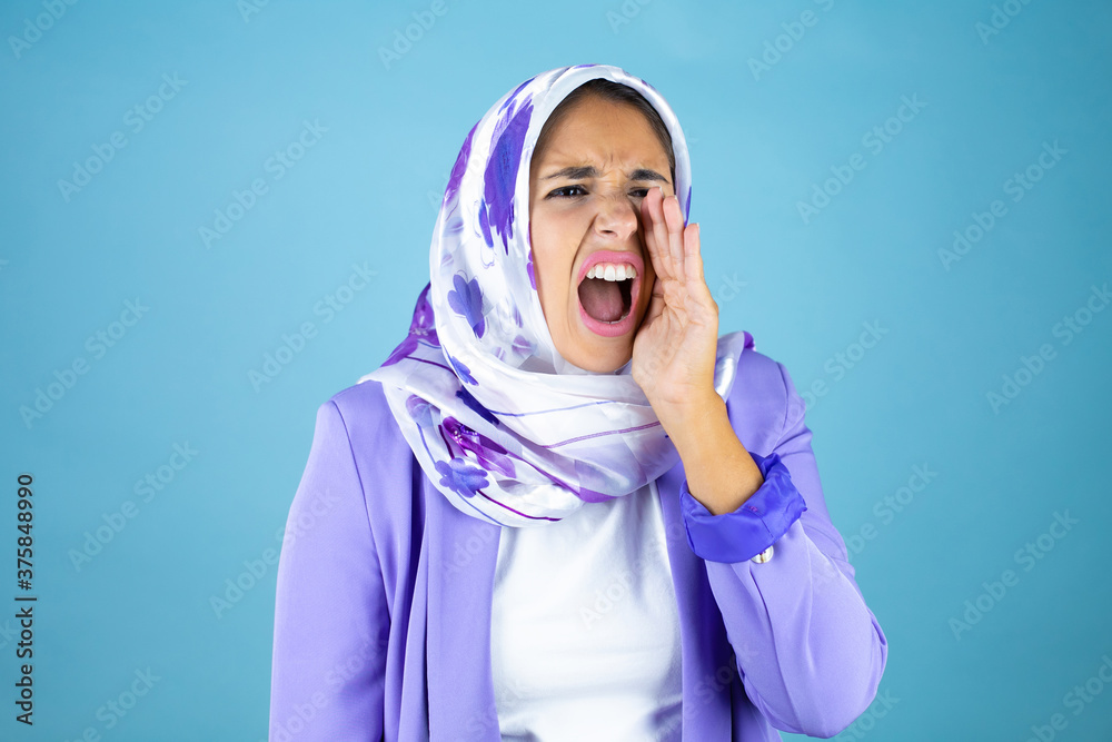 Young beautiful arab woman wearing islamic hijab over isolated blue background shouting and screaming loud to side with hand on mouth Young beautiful arab woman wearing islamic hijab over isolated blue background shouting and screaming loud to side with hand on mouth