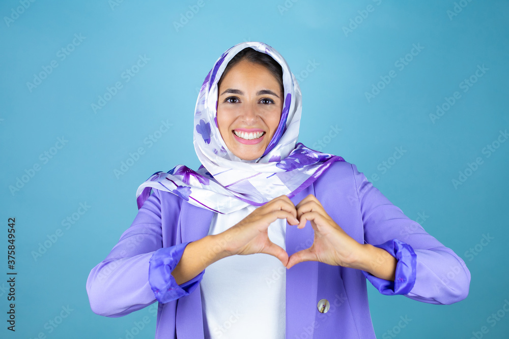 Young beautiful arab woman wearing islamic hijab over isolated blue background smiling in love showing heart symbol and shape with hands Young beautiful arab woman wearing islamic hijab over isolated blue background smiling in love showing heart symbol and shape with hands