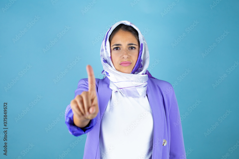 Young beautiful arab woman wearing islamic hijab over isolated blue background showing and pointing up with fingers number one while is serious Young beautiful arab woman wearing islamic hijab over isolated blue background showing and pointing up with fingers number one while is serious