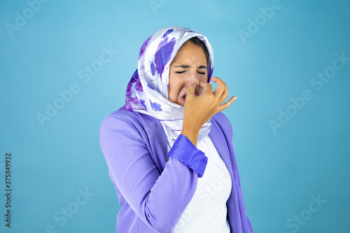 Young beautiful arab woman wearing islamic hijab over isolated blue background smelling something stinky and disgusting, intolerable smell, holding breath with fingers on nose © Irene