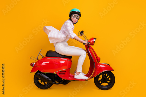 Side profile full length photo of cute student lady riding motorbike wind face excited high speed wear white striped shirt pants helmet glasses isolated yellow vivid color background © deagreez