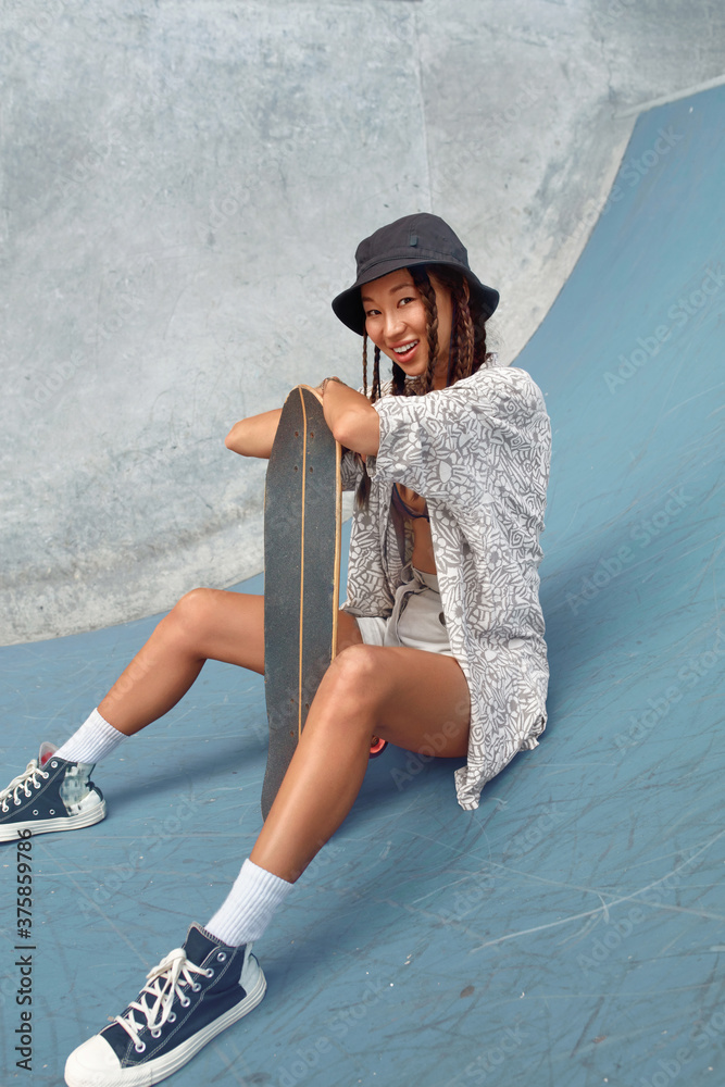 Foto de Skateboarding. Asian Girl In Urban Outfit With Skateboard Sitting  On Ramp Against Concrete Wall At Skatepark. City Sport As Hobby For Active  Teens. Skater Subculture And Summer Activity As Lifestyle.