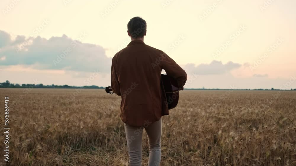 A back view of a young man is playing the guitar while walking in the field A back view of a young man is playing the guitar while walking in the field