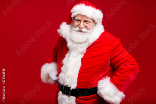 Photo of retired old man grey beard hands hips playful look self-assured ready long travel prepare sleigh wear santa costume belt coat spectacles headwear isolated red color background