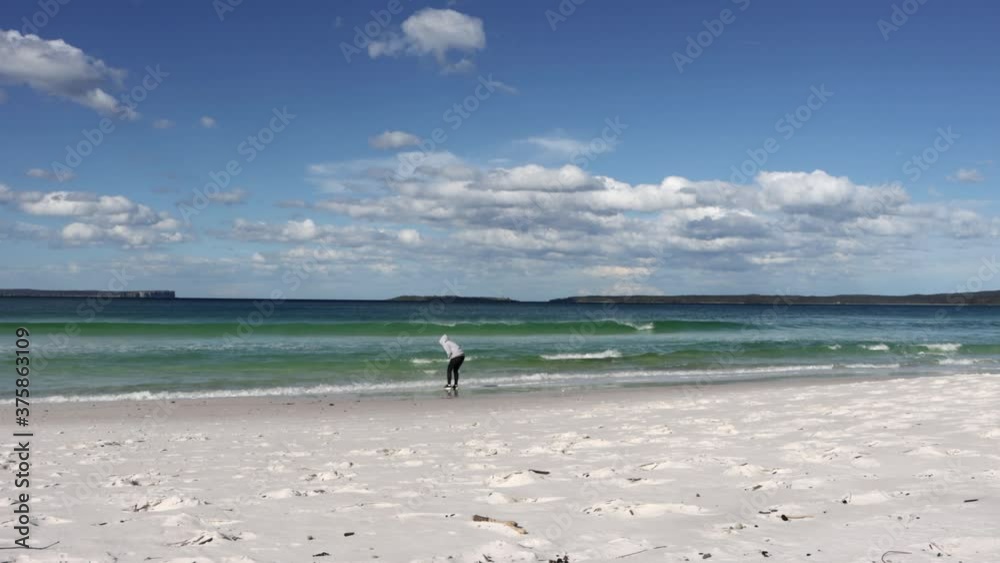 Hyams Beach in Jervis Bay with a woman in hoodie picking seashells from the sandy shore, Locked wide shot Hyams Beach in Jervis Bay with a woman in hoodie picking seashells from the sandy shore, Locked wide shot