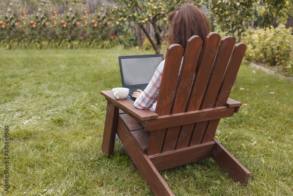 Fototapeta premium Young woman sitting in the garden in a chair and working remotely on a laptop on a background of grass and flowers. Rear view