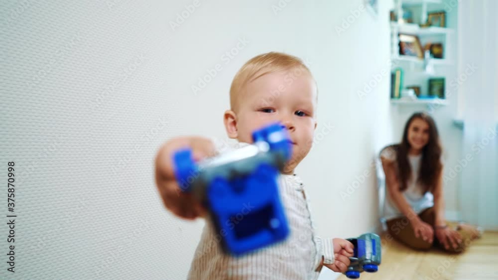 Little boy playing with toy cars at home, giving them to person behind camera, blurred mother sitting on floor on background. Kind child spending time at home. Concept of generosity Little boy playing with toy cars at home, giving them to person behind camera, blurred mother sitting on floor on background. Kind child spending time at home. Concept of generosity