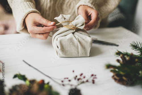 Female in cozy sweater preparing plastic free christmas present, zero waste. Hands wrapping stylish christmas gift in linen fabric on white rustic table with fir, pine cones, scissors, twine. photo