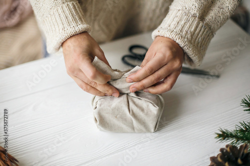 Woman in cozy sweater preparing plastic free christmas present, zero waste holidays. Female hands wrapping christmas gift in linen fabric on wooden table with green branch, pine cones, scissors. photo