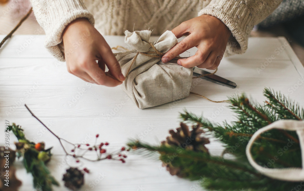 Female hands wrapping stylish christmas gift in linen fabric on white rustic table with green branch, pine cones, scissors and twine. Woman in cozy sweater preparing plastic free christmas present