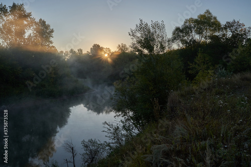 Fototapeta Naklejka Na Ścianę i Meble -  Autumn time. Dawn over the river in a misty, brooding haze. Beautiful view of the forest and river, covered with fog in the early morning. The sun's rays of light. September.