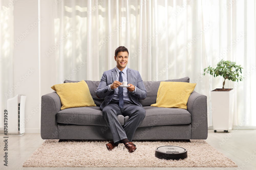 Robotic vacuum cleaner cleaning the floor and a young man in a suit sitting on a sofa
