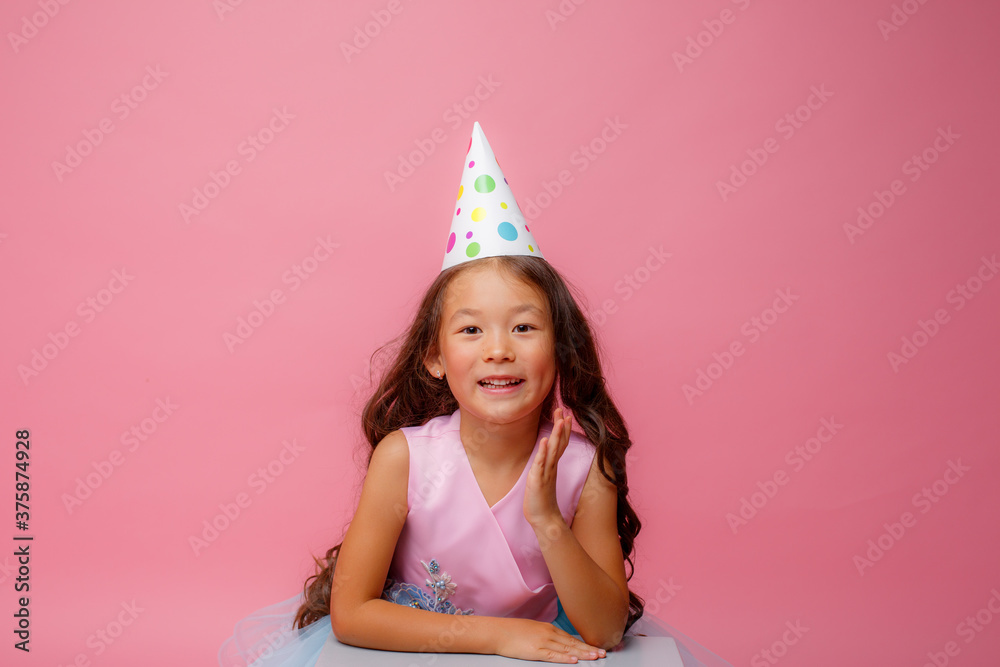 a little girl of Asian appearance with a cap on her head celebrates her birthday on a pink background