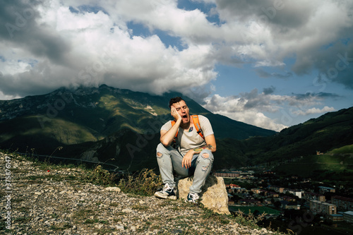 Tired traveling man resting on hill. Exhausted male tourist sitting on stone and sweetly yawning with closed eyes after active trekking in mountains