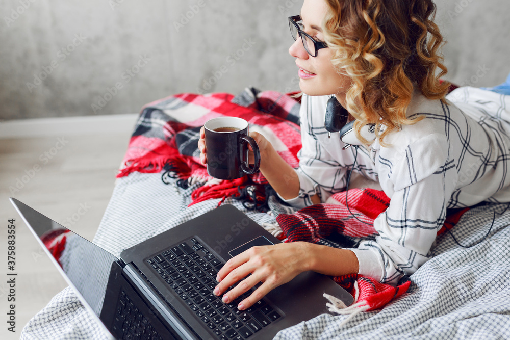 Stylish smart cheerful  woman in  eyeglasses using laptop , lying on cozy bed, drinking coffee.