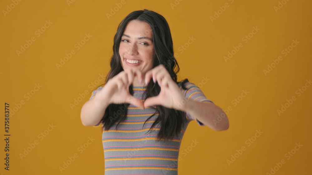A beautiful young woman is showing heart sign standing isolated over yellow background A beautiful young woman is showing heart sign standing isolated over yellow background